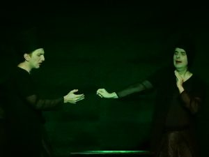 Zwei Schauspieler auf der Bühne in grünes Licht getaucht. Beide haben rot geschminkte Lippen und tragen eigenwillige Hauben. Sie geben sich die Hand