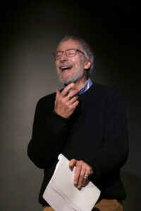 Wolfgang Keuter im Sprechtraining. Es steht auf schwarzer Bühne, hält einen Text in der Hand und lacht.