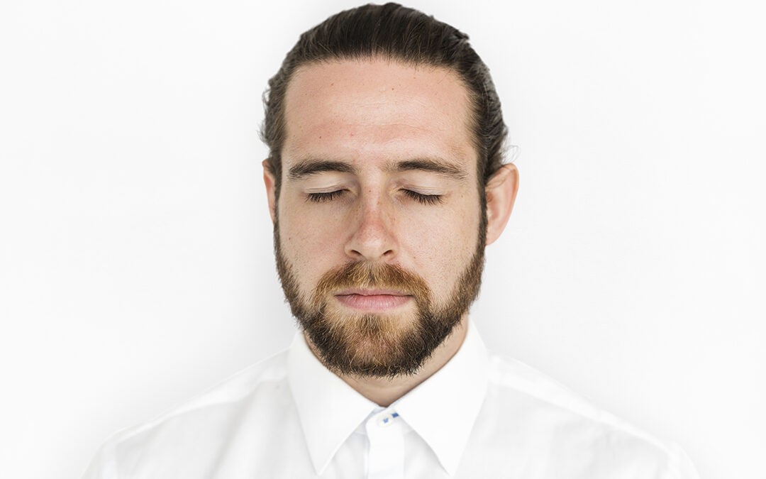 Ein junger Mann mit Bart steht vor weißem Hintergund mit geschlossenen Augen. Er trägt ein weißes Hemd.