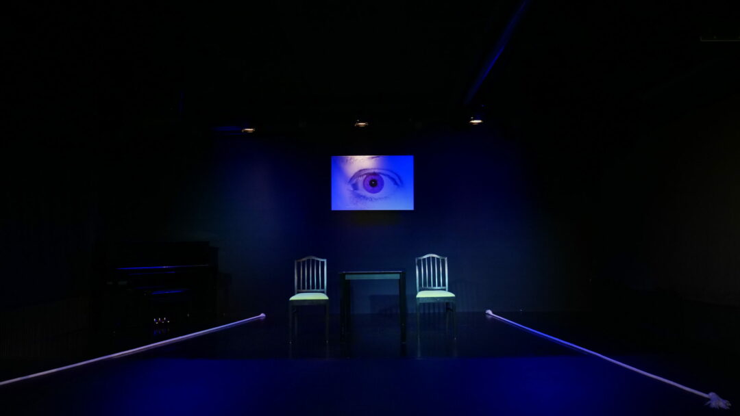 Auf schwarzer Bühne hängt in der Mitte ein geroßes Bild, welches ein auge zeigt. Davor stehen zwei schwarze Stühle mit grünem Polster und ein schwarzer Tisch in der Mitte