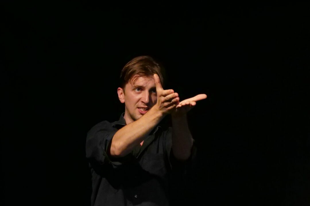 Junger Mann auf schwarzer Bühne zeigt eine Gebärde im Stile von Slow Acting